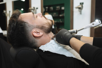 Close-up of hairdresser work for attractive young man at barbershop. Styling beard, shaving it off with a straight razor