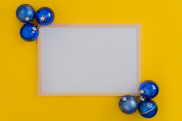 wooden photo frame on a yellow background with blue Christmas balls in the corners. Color contrast, horizontal photo. Idea - New Year's photo frame, template for advertising, congratulations, text.