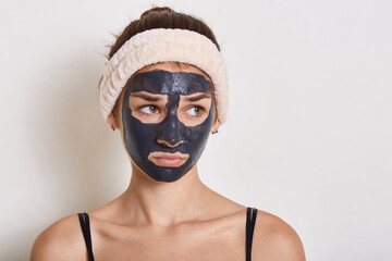 Sad teenage girl with face mask looking aside, expressing sadness, wearing sleeveless t shirt and hairband, stands against white background, cosmetology.
