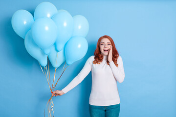 Surprised red haired female with widely opened mouth, touching her cheek with palm, wearing casual...