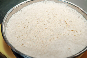 Pulling the plastic wrap off the raised bread dough in a metal mixing bowl. The gluten is stretched, showing that the raw flour mixture can be formed into a loaf and baked.