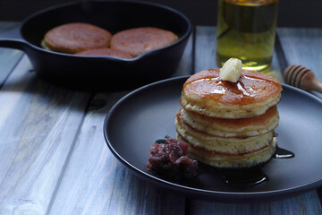 stack of butter pancakes with honey and red bean paste in the black round dish on the wooden table with background of bakery pan and olive oil in the kitchen of the room during tea time with family
