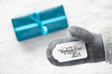 Label With German Calligraphy Frohe Weihnachten Und Ein Glueckliches 2021 Means Merry Christmas And A Happy 2021. Gray Glove With Turquoise Gift And Snow Background