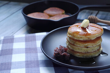 stack of butter pancakes with honey and red bean paste in the black round dish on the wooden table with background of bakery pan and olive oil in the kitchen of the room during tea time with family