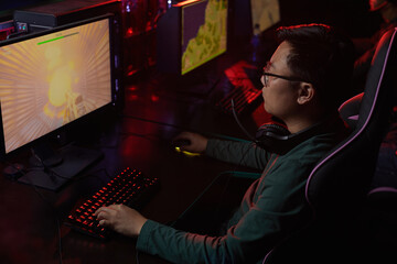 Asian young gamer in eyeglasses looking at computer monitor he concentrating on his game in dark computer club