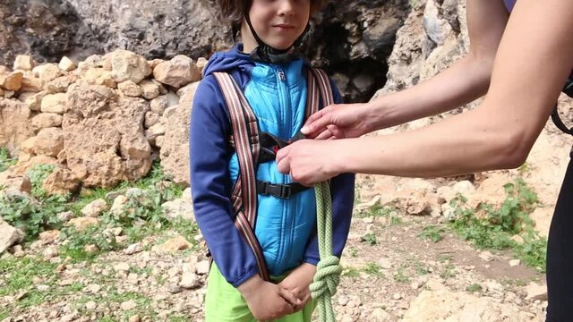 The instructor teaches the child to use climbing equipment, The boy is preparing to climb a rock, A woman shows a child how to use a carabiner for belaying, Mother ties the rope to the safety system.