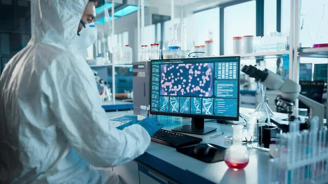 Medical Laboratory: Microbiology Scientist Wearing Sterile Coverall, Face Shield and Mask Works on Computer Developing Vaccine, Drugs and Antibiotics. Screen Shows High-Tech Concept for DNA research