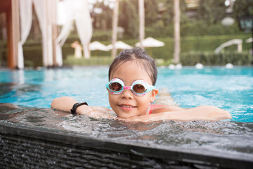 smiling child wearing swimming glasses in swimming pool. little girl playing in outdoor swimming pool on summer vacation on tropical beach island. child learning to swim in pool of luxury resort.