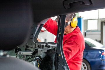 Expert senior mechanic with yellow headphones, repairing a car after a street crash. Details and focus on metal parts and electric wires. Blurred background, natural light. Occupation and job concept.