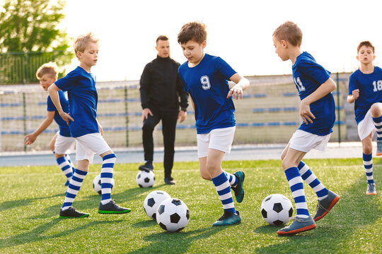 Happy boys on a sports training class with young coach. School age children making sport. Kids kicking soccer ball on summer football camp