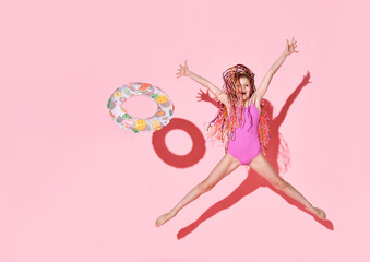 Happy girl with bright cornrows in one-piece rose swimsuit jumping and screaming full of joy, throwing rubber ring with fruit pattern