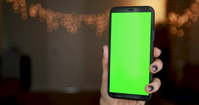 Young woman hand with silver manicure holds black smartphone with green screen against lights of orange colour closeup