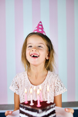 Little blonde girl smiling in birthday pink cap, a chocolate birthday cake with candles.