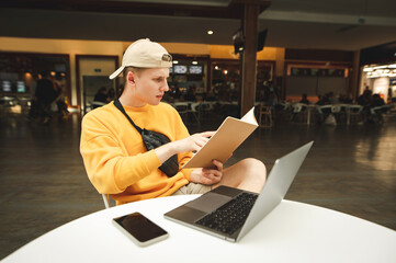 Portrait of handsome student boy in casual clothes sitting with laptop on desk and book in hands in cafe, reading literature with serious face. Young male student studying on break in cafe.