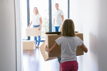 Fototapeta na wymiar Back view of blonde girl carrying cardboard box and entering into room of new apartment or house. Parents in jeans standing inside with carton boxes and smiling. Relocation and moving day concept