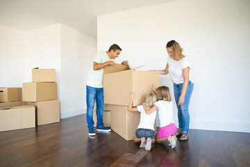 Parents and two daughters having fun while opening boxes and unpacking things in their new empty flat. Girls sitting on floor. Real estate purchase concept