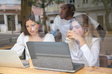 Content multiethnic designers talking about design indoors behind window. Caucasian ladies drinking coffee and sitting. African American man looking at laptop. Communication and teamwork concept