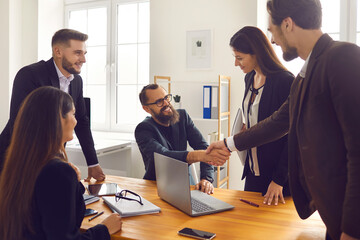 Smiling business partners shaking hands after successful negotiations with colleagues in office