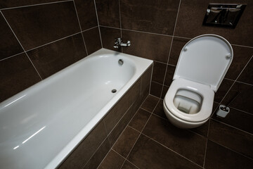 Detail of a small bathroom with a bathtub and a toilet next to each other. The toilet is modern with an open seat. Paving and tiling have the texture of a concrete trowel in shades of brown.