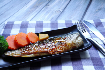  grilled mackerel in the black plate with carrot and broccoli with background of blue and white towel and wooden table in the Japanese restaurant during dinner with family in the party