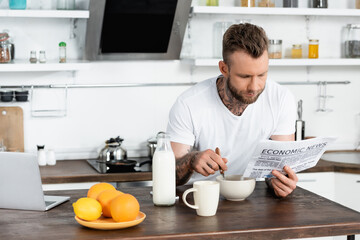 tattooed man in white t-shirt having breakfast in kitchen and reading newspaper