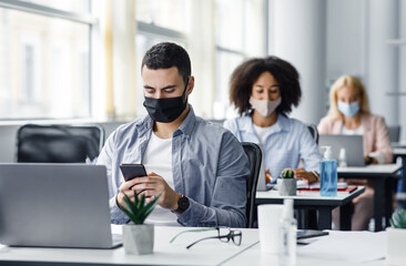Modern gadgets for working with clients remotely from office after quarantine. Arabic young guy in protective mask typing on smartphone at table with laptop in interior