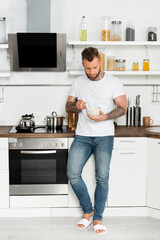 young man in white t-shirt and jeans holing bowl with breakfast while standing in kitchen