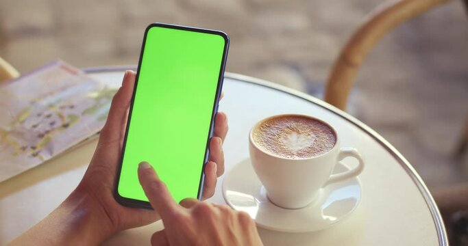 Crop view of female person holding mobilephone and pressing on greenscreen while sitting at table with coffee cup and map on it. Concept of mockup and chroma key.