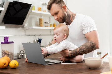 tattooed man holding infant son touching laptop while sitting on kitchen table