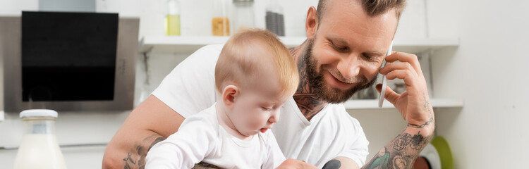 horizontal image of young man talking on smartphone in kitchen during breakfast with infant son