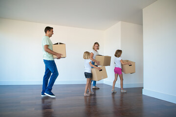 Happy family carrying cardboard boxes from empty room. Two little daughters helping parents with relocation in new apartment or house. Mortgage, relocation and moving day concept