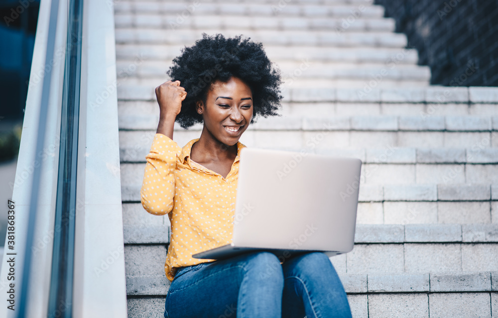 Wall mural A young beautiful curly woman sitting on the stairs, looking at her laptop and showing a symbol of success with her hand