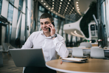 Businessman talking on smartphone while sitting with laptop in cafeteria