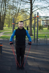 Young sporty bearded man doing exercises with rubber band outdoor. Fitness guy training with elastic band at sports groung. Athlete doing dynamic workout.