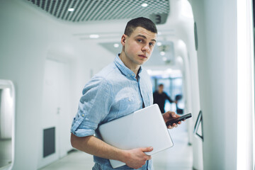 Serious man with laptop using smartphone in contemporary hallway