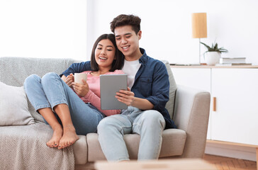 Asian Couple Using Digital Tablet Sitting On Sofa At Home