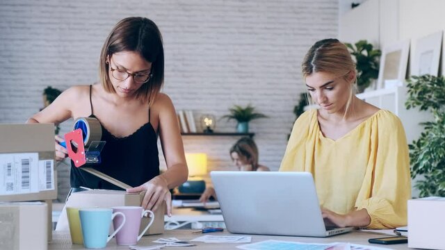 Video of two beautiful freelance business women seller checking product order with computer while packing and sealing cardboard boxes for delivery products in their startup small business. 