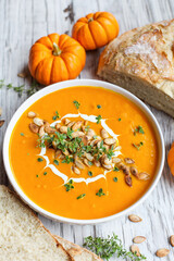 Fresh pumpkin soup garnished with sour cream, toasted pumpkin seeds and thyme with homemade artisan bread over a white wood background. Selective focus with blurred background.