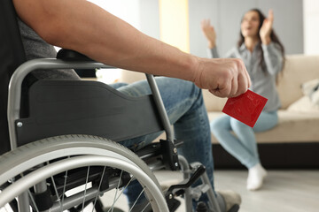 Obraz premium Man sits in wheelchair and holds condom in his hands opposite woman sits and happily raises his hands up. Intimate life with disability concept