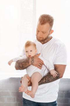 Young Bearded Man In White T-shirt Holding Infant Child In Baby Romper