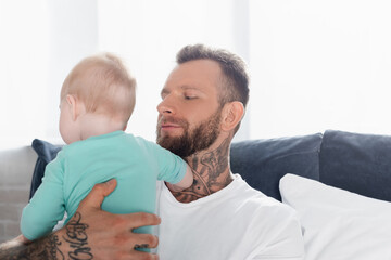 bearded, tattooed man holding infant child in bedroom