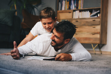 Young father lying down on the floor and reading book to his beloved son.