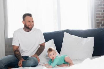 excited young man in white t-shirt and jeans sitting near baby boy crawling on bed with open mouth