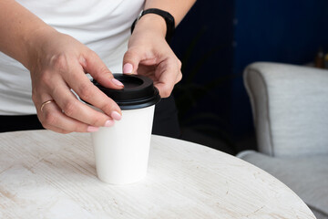 Female hands put a black lid on a white paper cup with hot coffee or tea to be taken outside, close-up