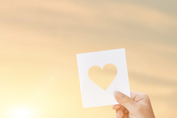 Silhouette of hand is raising paper with shape heart at sunset background.