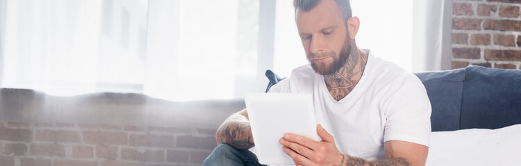 horizontal image of bearded tattooed man using digital tablet in bedroom