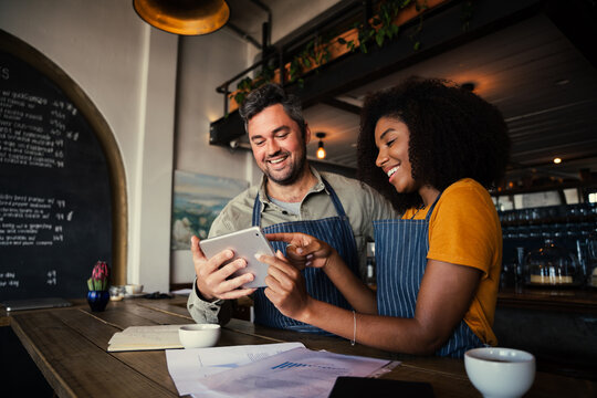 Manager and waitress laughing at designs on digital tablet standing with hot coffee in cafe 