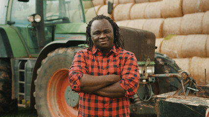 Young african farmer with crossed hands over the chest in front of tractor and haystack. High...