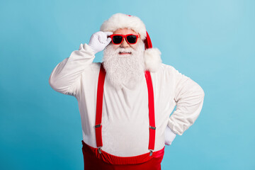 Fototapeta na wymiar Photo of pensioner old man grey beard hold specs hand hip self-assured prepare check sledge wear santa costume red suspenders spectacles white gloves headwear isolated blue color background