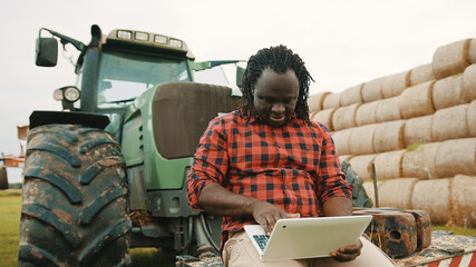 Fototapeta na wymiar Young african farmer using tablet while sitting on green tractor. Haystack in the background. High quality photo
